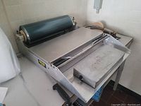 Stainless steel butcher wrapping station with a mounted roll of dark plastic wrapping film. Side handle and cutting bar visible.