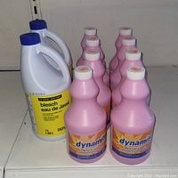 Photo showing two large bottles of bleach and eight bottles of Dynamite pink container cleaners, all placed on a flat surface.