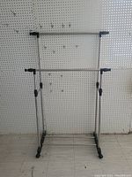 Front view of metal clothes rack showing two extendable horizontal bars and wheeled base with one wheel missing.