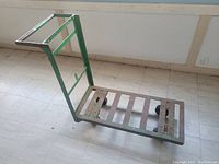 Green metal dolly cart with worn paint and metal slat platform, photographed from side angle showing the wheels and handle.