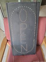 Photo showing front view of the black rectangular open sign with LED bulbs spelling OPEN arranged vertically and an oval border of bulbs. Sign has a hanging chain and slight crazing visible on surface.