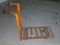 Side view of the orange metal dolly cart showing the handle and base with wheels on tiled floor.