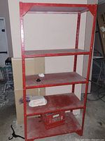 Front view of red metal shelving rack showing four dusty shelves, with some miscellaneous items placed on lower shelves.