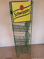 Front view showing green metal wire rack with multiple angled shelves and yellow Schweppes sign at top.