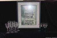 Photo showing framed print centered with eight clear crystal wine glasses on the left and four green stem wine glasses on the right against a black backdrop.