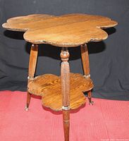 Full view of antique cloverleaf table showing wooden cloverleaf-shaped tabletop and lower shelf, turned legs with claw glass ball feet, standing on red carpet against black backdrop.