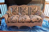 Full front view of antique wicker sofa with floral-patterned cushions placed on a blue tarp. Cushion fabric shows visible wear with fading and some damage on throw pillows. Paint on frame is chipping.