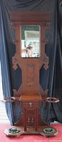 Full front view of antique wooden hall tree showing central mirror, upper carved details, drawer with knob, and cast iron umbrella holders attached at base on wooden platform.