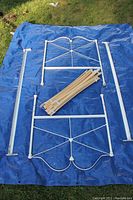 Overview of all white wrought iron bed frame components laid on blue tarp: headboard, footboard, side rails, wooden slats base, and hardware.