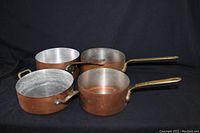 Four copper pots with brass handles laid out on black background, showing different sizes and condition.