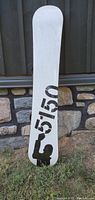 Base of the snowboard showing white surface with black '5150' text and figure graphic, placed vertically against stone wall on grass.