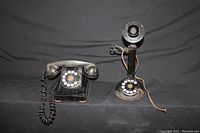 Front and side view of two antique rotary telephones on black backdrop, candlestick phone missing earpiece, desk phone with coiled cord