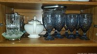 Wood cabinet shelf containing ten blue frosted etched wine glasses with floral patterns on the right, clear glass relish tray on the left, and white ceramic JA Browne teapot with gold accents center-left.