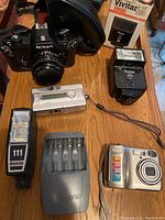 Photo showing full lot including Nikon FM film camera with case, Vivitar 2600 and Dimension 111 flashes, silver digital camera and charging devices on wooden table.