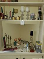 Shelf with numerous candles of different colors and various candle holders made of glass, metal, and ceramic.