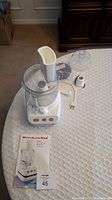 Full picture of assembled KitchenAid food processor with lid, bowl, and base on table along with user manual and lot tag