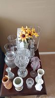 Photo showing entire collection of various vases on dark wooden table including clear glass, frosted, pink ruffled edge, and ceramic vases. Some vases contain artificial yellow flowers.