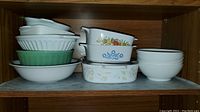 Wooden shelf with multiple stacked bowls and casserole dishes, including CorningWare Blue Cornflower pattern dishes and plain white bowls