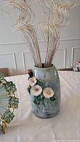 Tall ceramic vase with white flowers and green leaves attached, glazed in greenish hues, placed on a white cloth.