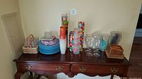 Overall view of collection on wooden table showing stacked blue melamine plates and bowls, plastic tumblers, colorful polka dot melamine glasses, clear plastic pitchers, wine glasses, and wicker basket accessories.