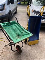 Green fabric multipurpose garden cart with black metal frame and two small wheels seen next to blue wheelbarrow/cart