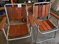Pair of wooden folding outdoor chairs with aluminum frames and brown wood slats for seat and backrest, along with brown plastic armrests, shown from front in garage setting.