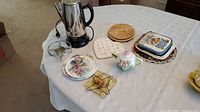 Photo of Cuisinart electric percolator, porcelain teapot, woven coaster trays, and ceramic serving plates on table