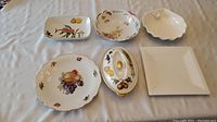 Full set of six porcelain serving dishes in assorted shapes with fruit and floral designs laid on fabric.