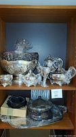Two shelf view of multiple silver tea set items including teapot, coffeepot, bowls and trays arranged inside a wooden shelving unit.