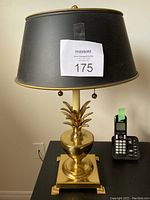 Full view of the brass pineapple table lamp with black lampshade and brass finial, showing the lamp on a black table next to a cordless phone.