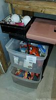 Pink plastic toolbox sitting on top of two clear plastic bins filled with miscellaneous items, and a small black case containing soldering supplies.