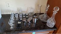 Wide shot showing two crystal cut decanters with stoppers, an ice bucket, assorted wine glasses including frosted shot glasses, and black wine accessory tool on a dark counter.