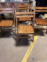 Four wooden folding chairs with slatted seats and backs in medium brown wood with brass hardware placed on a concrete floor.