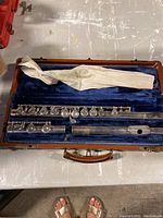 Full view of the silver flute disassembled into three parts, inside its blue velvet-lined case with a brown handle and white fabric sleeve.