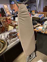 Front view of ironing board covered with beige padded cover, standing upright; set among various other household items.