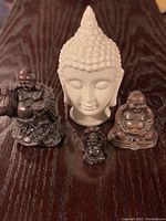 All four Buddha figurines including one large white ceramic Buddha head and three small dark metal laughing Buddhas displayed on a dark wooden surface.
