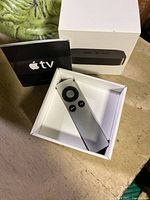 Apple TV remote control in box beside user manual and outer box on table.