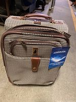 Front view of the London Fog carry-on suitcase showing the houndstooth fabric pattern, front zippered pocket with leather trimmed button flap, top handle, and attached manufacturer tag.