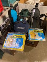 Photo showing three canteens, two green propane camping lanterns, and two yellow vinyl ponchos sealed in original packaging on a small table.