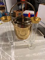 Front view of solid brass ice bucket with hinged lid and two champagne flutes with gold rims, placed on a flat surface.