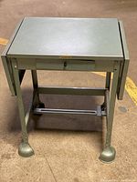 Front view of the metal typewriter cart showing the main top surface, drawer with pull, and legs with feet.