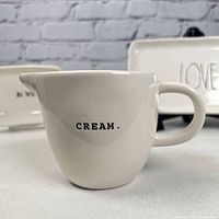 White ceramic cream pitcher with black 'CREAM.' label, handle and spout visible.