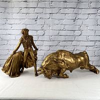 Full view of bullfighter and bull statues in metallic gold finish against a white brick background.