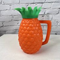Front and side views of the vintage orange Minerware plastic pitcher shaped like a pineapple with a green leafy top lid and an angular handle.