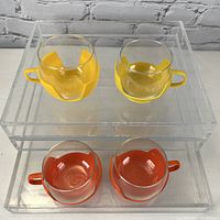 Four glass coffee mugs with yellow and orange plastic holders and handles, displayed on clear acrylic risers against a white brick background.