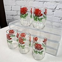 Five clear glass juice glasses decorated with red rose patterns, green leaves, and white picket fence motifs. Shown on a clear acrylic riser against a white brick background.