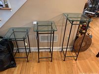 Three tables of varying heights with dark metal bases and ridged square glass tops placed side by side against a wall on a wood floor.