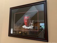 Framed art print featuring a red vase, glass, and white doily with dark background and reflection of room.
