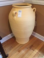 Full view of the tall beige clay jug, positioned on wooden floor near corner wall, showing overall shape and handles.