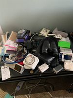 Photo showing a surface cluttered with various items including a wrist blood pressure monitor, binoculars, cords, a shot glass, and hosiery.